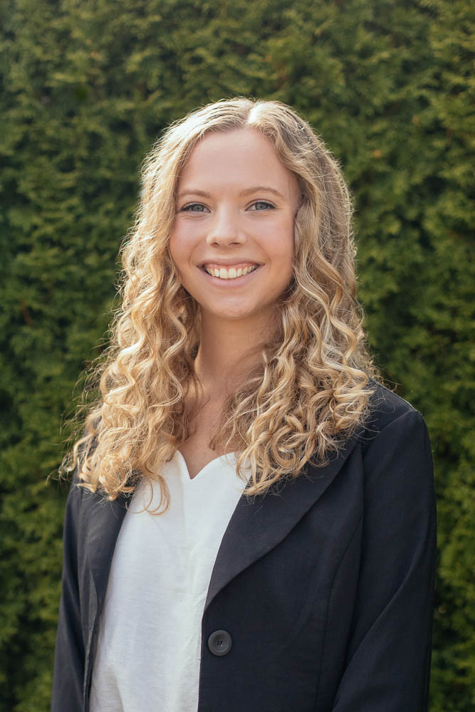 Amy, sterile tech assistant with long, curly blonde hair wears a white shirt and a black blazer, standing in front of green foliage. The outdoor setting and soft lighting create a natural and relaxed atmosphere.