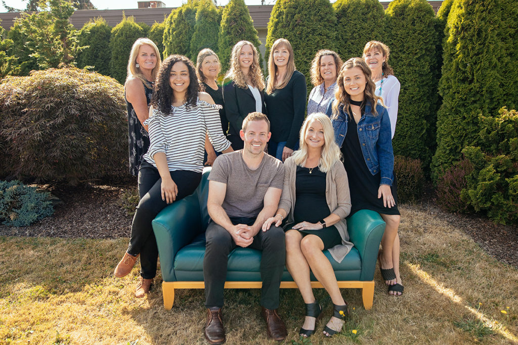 Ten people pose outdoors for a group photo, with eight standing in the background and two seated on a green couch in the foreground. The setting includes tall green shrubs and grass, with individuals dressed in a mix of casual and semi-formal attire.