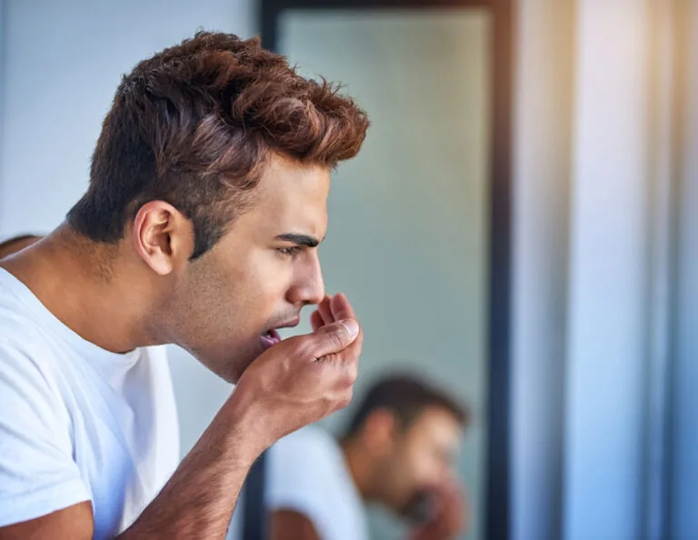 Person in a white shirt leaning towards a mirror, cupping a hand near the mouth, possibly smelling their breath.