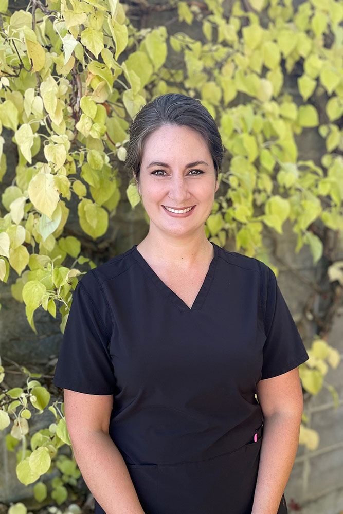 Headshot of Anna wearing a black scrub while her hands place in the front. A wall with vibrant green and yellow leaves and a serene atmosphere.