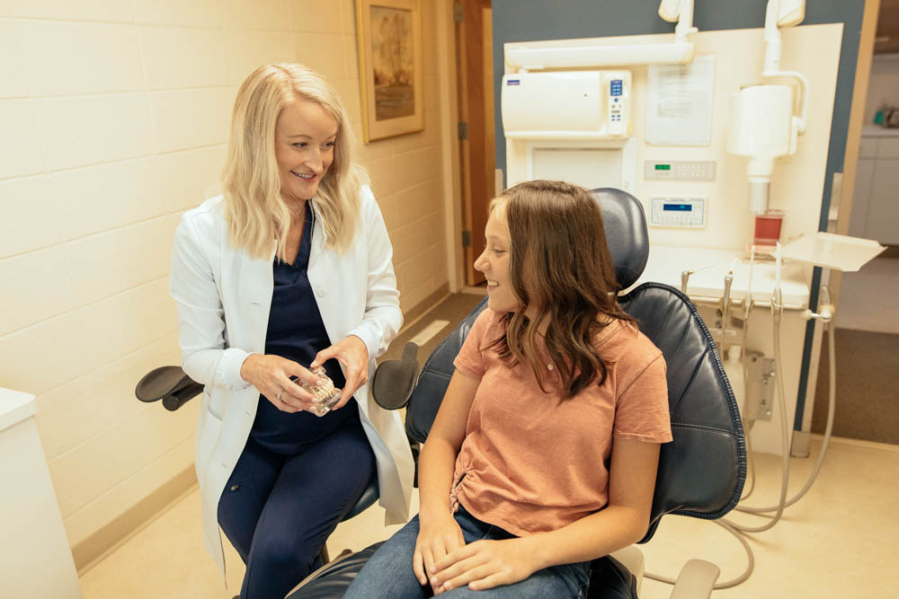 Dr. Whitney Forsythe holds a model of teeth while speaking to a dental patient who is seated in an exam chair