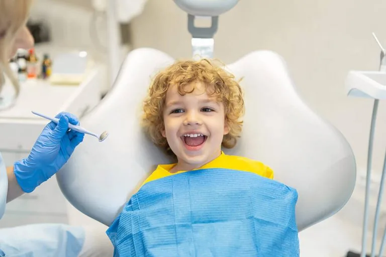 A child in a dental chair wearing a blue dental bib, with a dental professional holding a dental mirror.